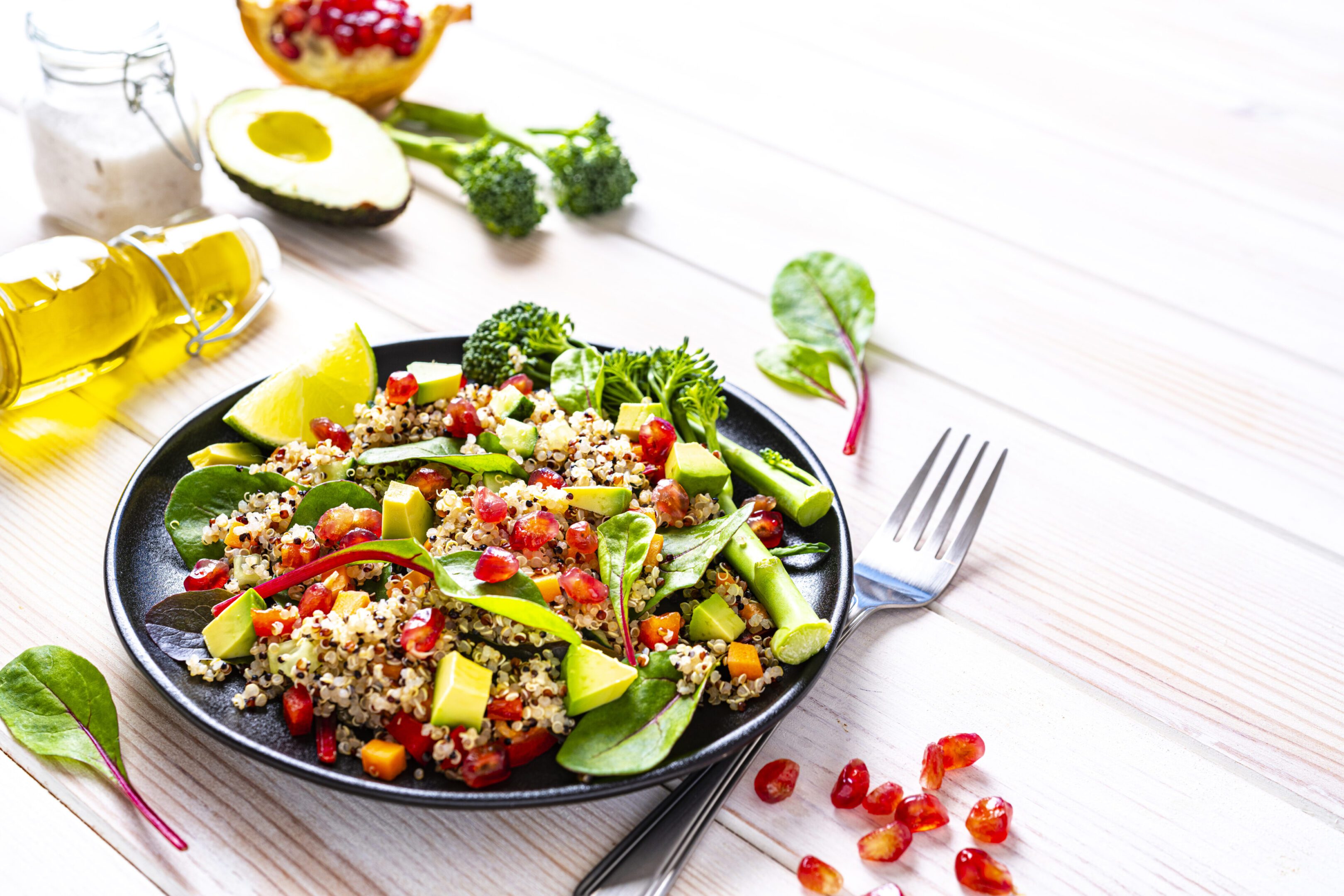 Vegan food backgrounds: healthy fresh multicolored quinoa salad plate shot on white table. The salad includes chopped carrot, avocado, pomegranate, broccoli, red chard leaves and tomato. Ingredients are around the plate. The composition is at the left of an horizontal frame leaving useful copy space for text and/or logo at the right. High resolution 42Mp studio digital capture taken with SONY A7rII and Zeiss Batis 40mm F2.0 CF lens
