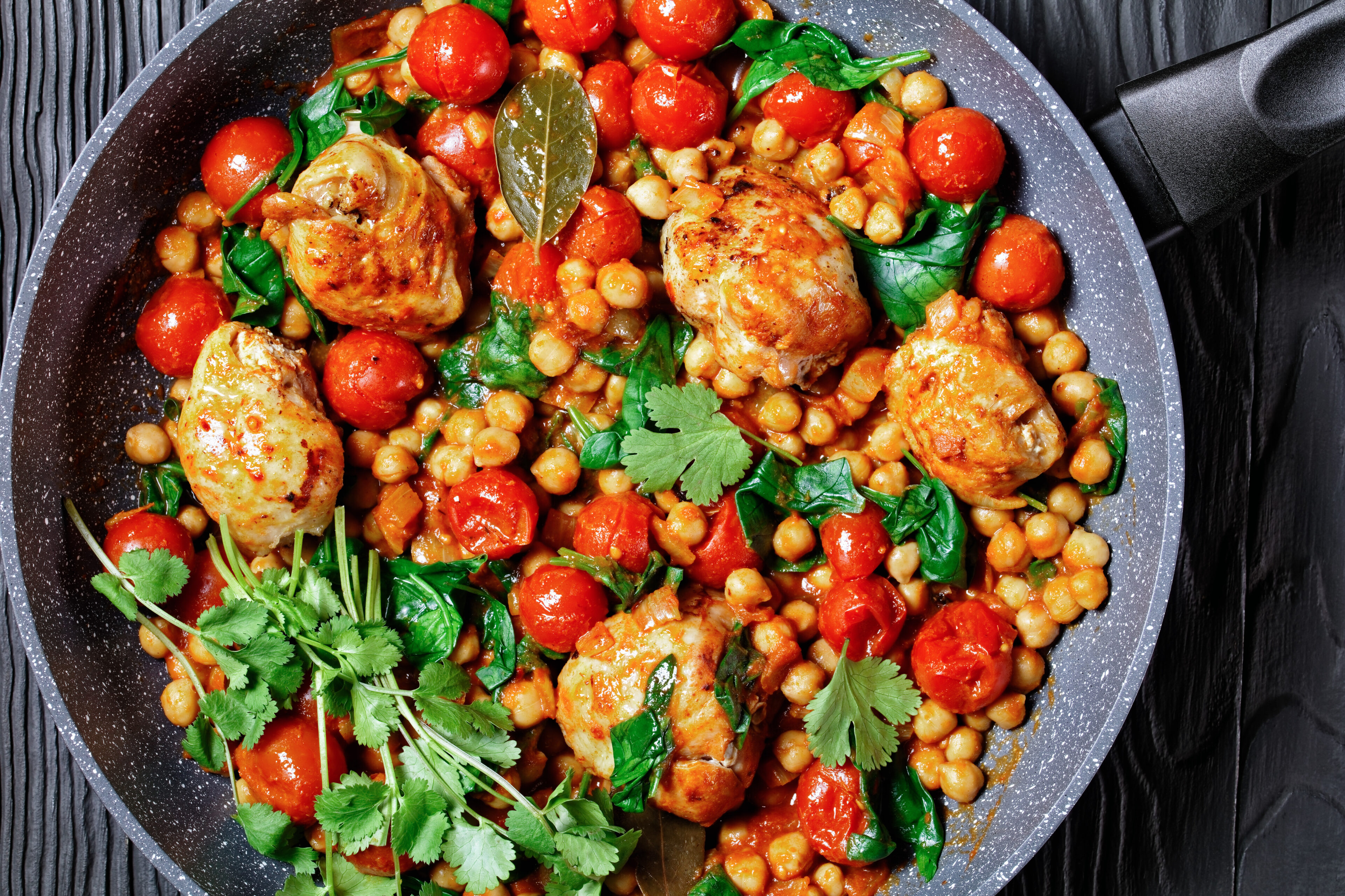 Indian cuisine: chickpea and chicken curry of boneless chicken thighs with cherry tomatoes and spinach served with fresh cilantro on a frying pan on a black wooden background, top view, close-up