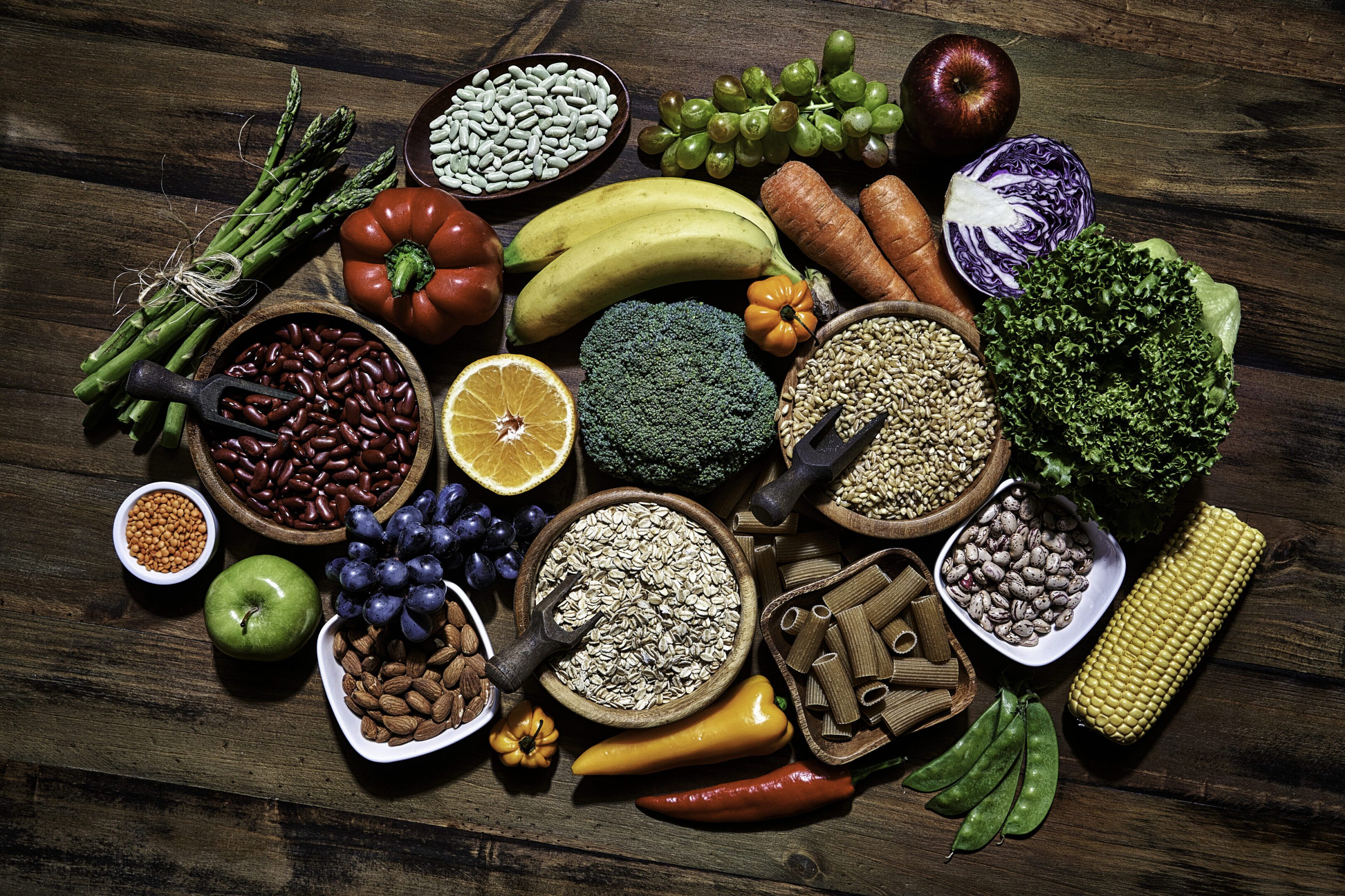 Dietary and healthy food themes. Table top view of fresh vegetables and legumes on rustic wooden table. Food is rich of fiber ideal for dieting and healthy eating. Includes corn, avocado, broccoli, orange fruit, grapes, bell pepper, lettuce, banana, apple  almonds and wholegrain pasta.