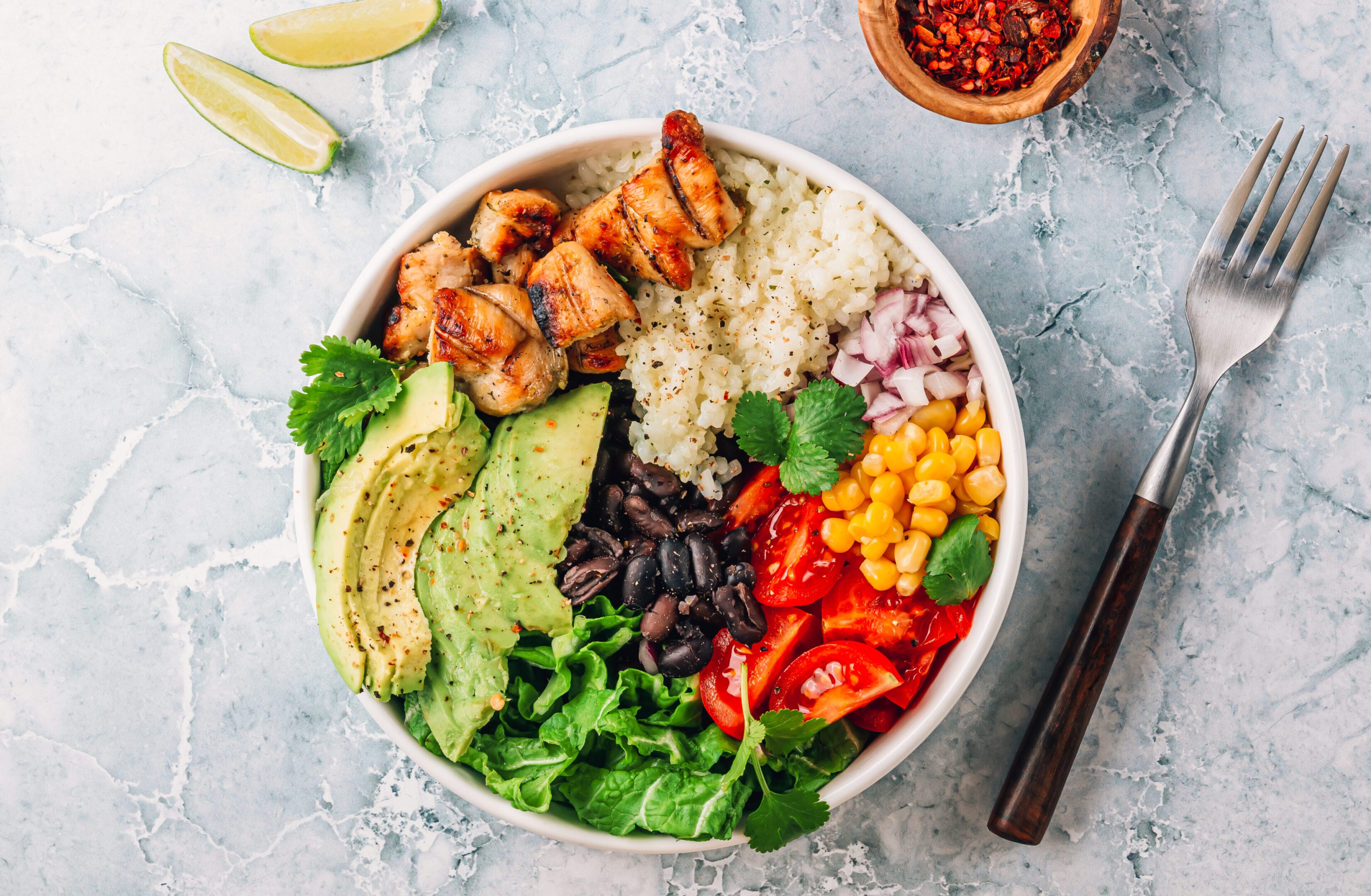 Mexican chicken burrito bowl with rice, beans, tomato, avocado,corn and lettuce over gray marble background. Mexican cuisine. Healthy food concept. Top view